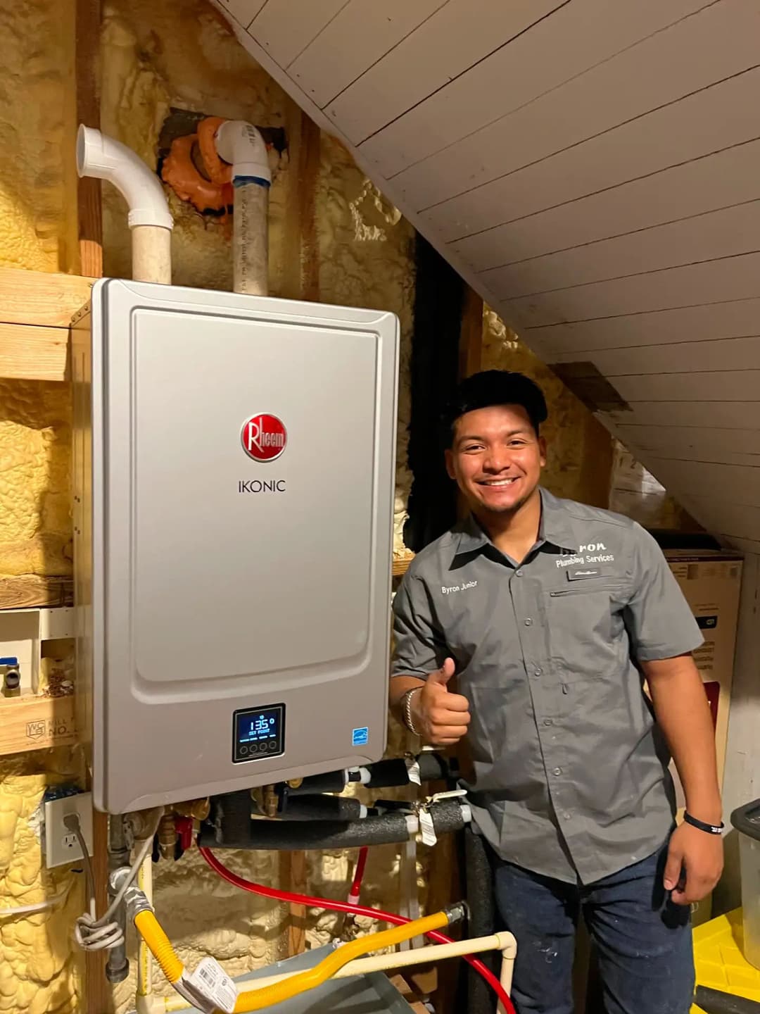 Technician with Rheem Ikonic water heater in an insulated attic setting, giving a thumbs up.