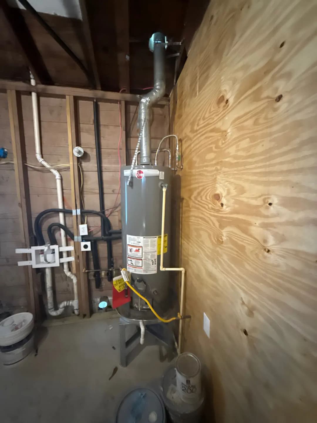 Residential water heater installed in a basement with exposed plumbing and wood paneling.