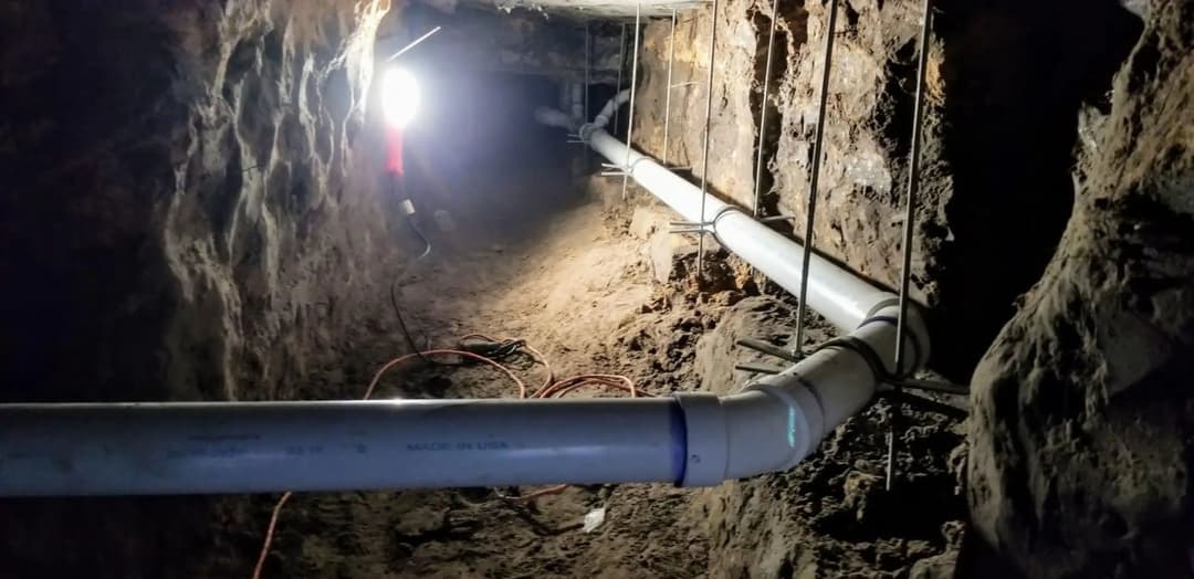 Underground tunnel with PVC pipes, dimly lit by a light source, surrounded by rocky terrain.