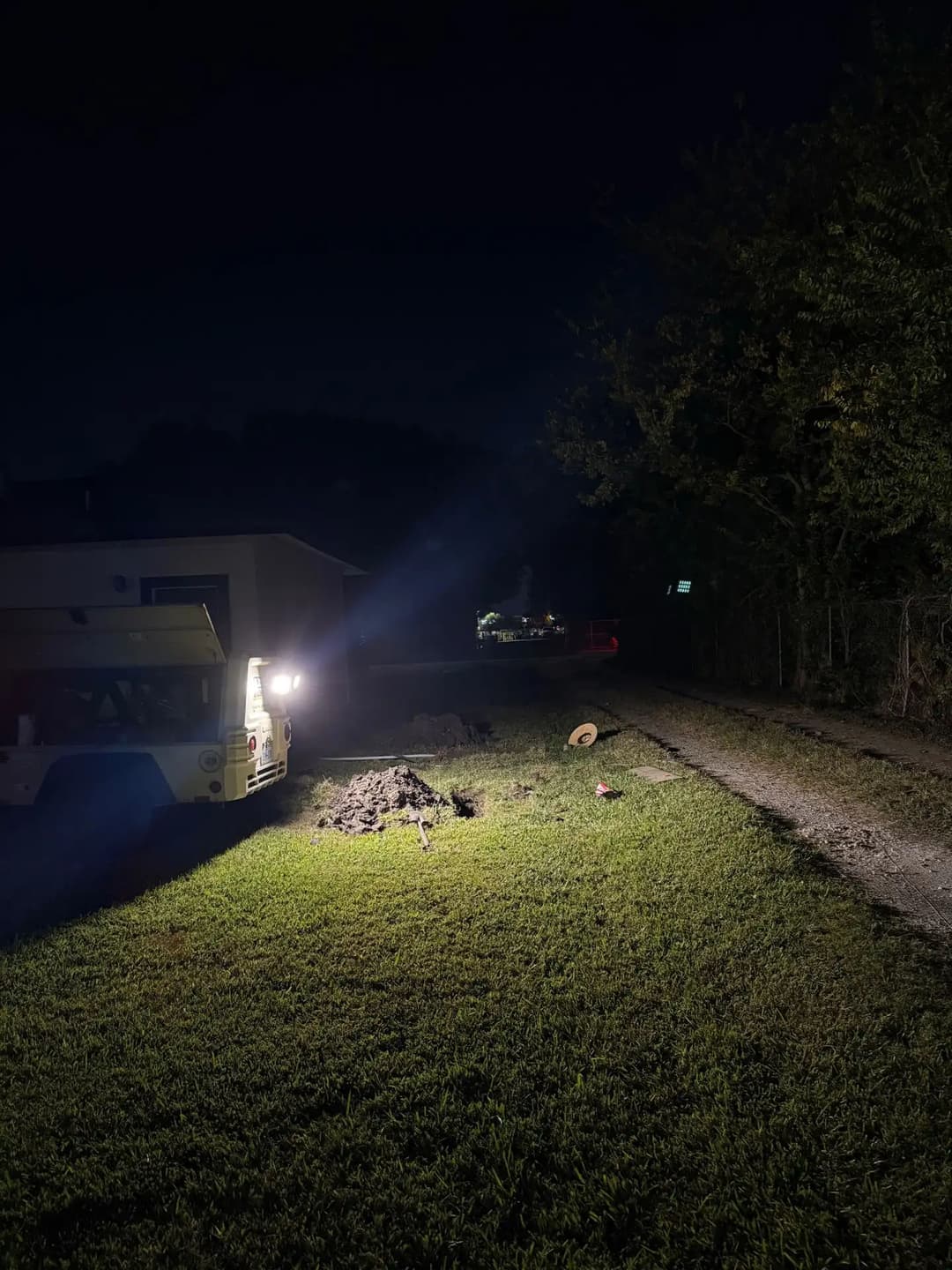 Nighttime scene with a lit path, excavated earth, and a distant vehicle on a road.