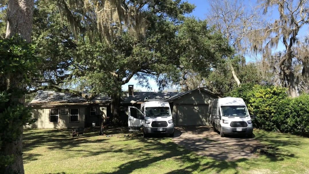 Two white vans parked in front of a house under a large tree with Spanish moss.
